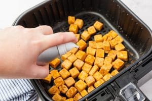 hand spraying air fryer tofu in black air fryer basket