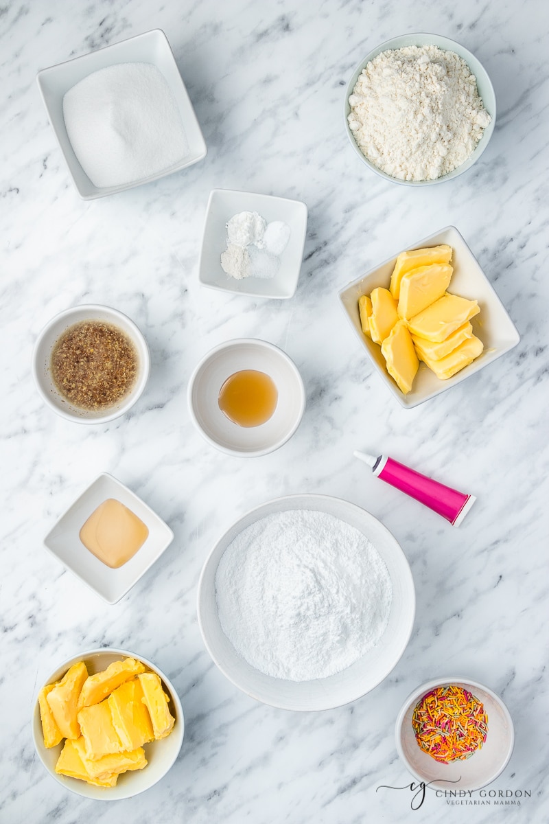 White bowls of flour, powdered sugar, vegan butter, sprinkles, vanilla extract, salt, and sugar