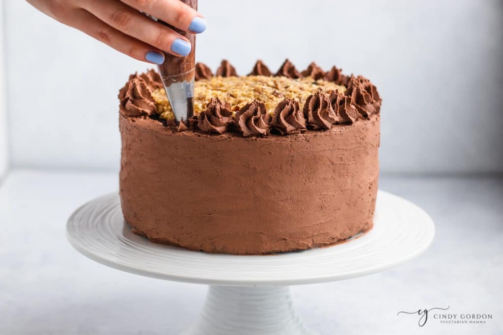 a german chocolate cake on a cake stand. A hand is adding icing stars to the top of it