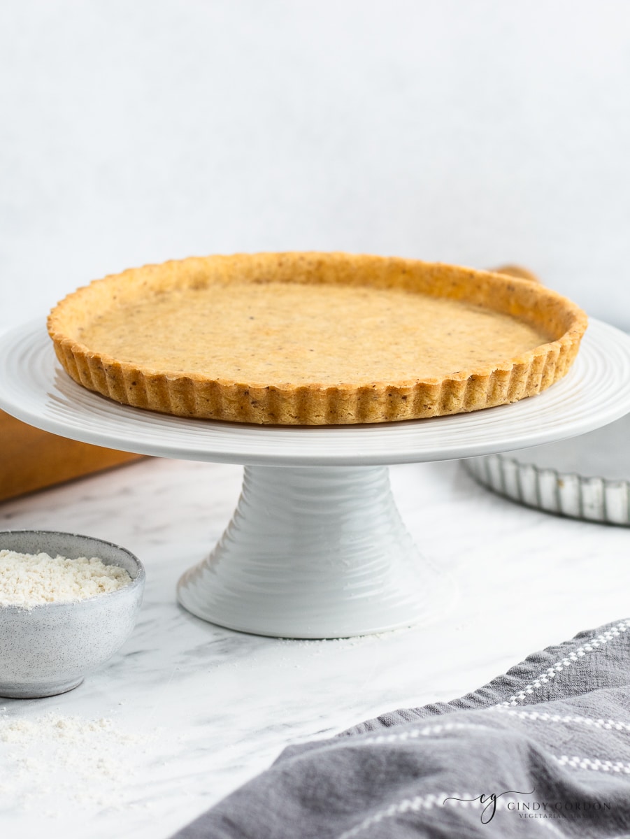 A shallow pie crust on a white pie stand nest to a bowl of flower and a metal pie plate