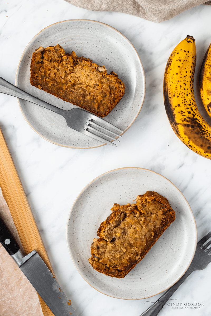 2 slices of banana bread on gray round plates next to bananas