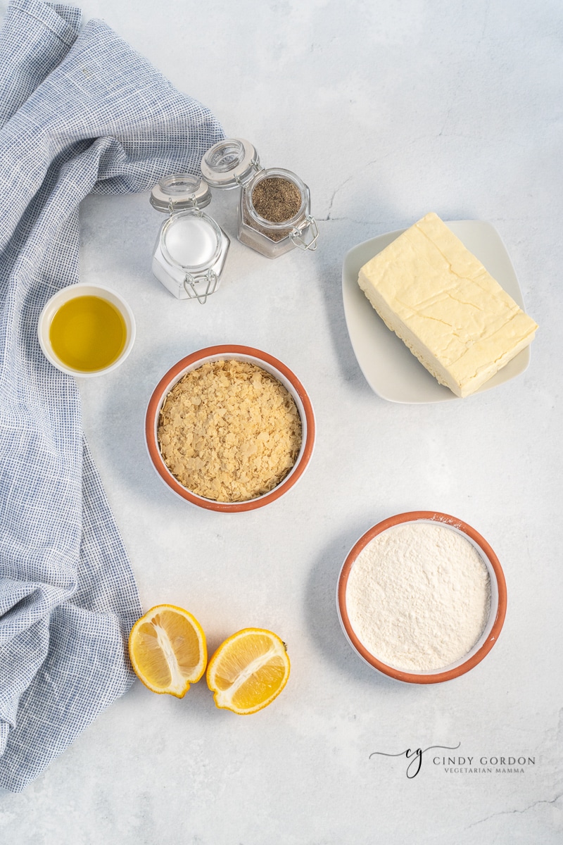 Tofu on a plate surrounded by lemons, nutritional yeast, oil, flour, salt, and pepper