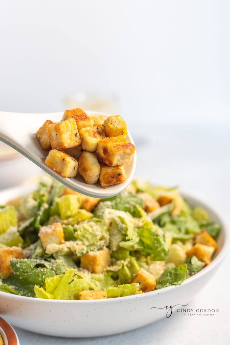 A spoonful of tofu croutons over a bowl of Caesar salad