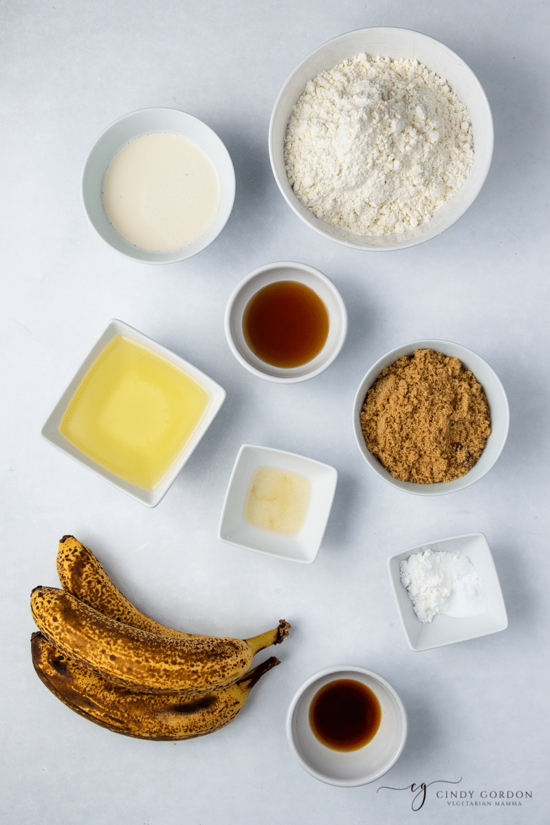 Bananas next to bowls of flour, brown sugar, milk, vanilla, oil, sugar, and apple cider vinegar