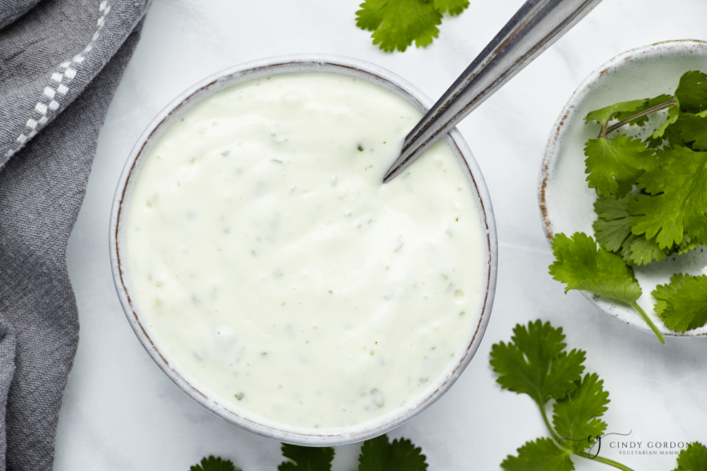 white bowl on a white background, filled with white sauce. garlic, cilantro, lime cut open laying around the bowl