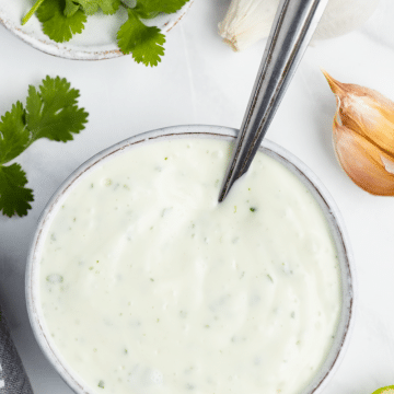 white bowl on a white background, filled with white sauce. garlic, cilantro, lime cut open laying around the bowl