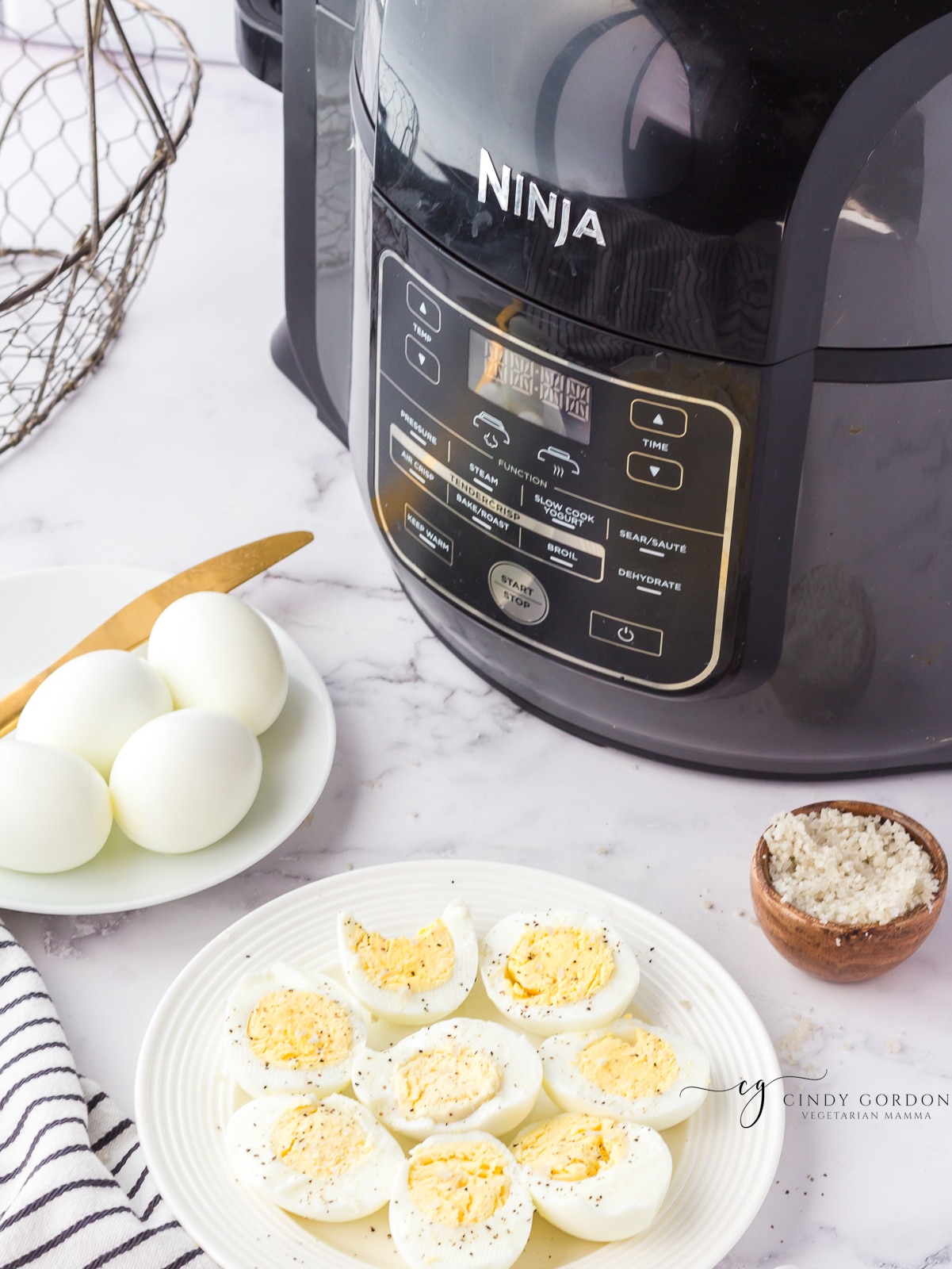 A ninja foodi multicooker next to two plates of hard boiled eggs. one plate has halved eggs with salt and pepper and one has whole eggs. A bowl of coarse salt is also on the counter. 
