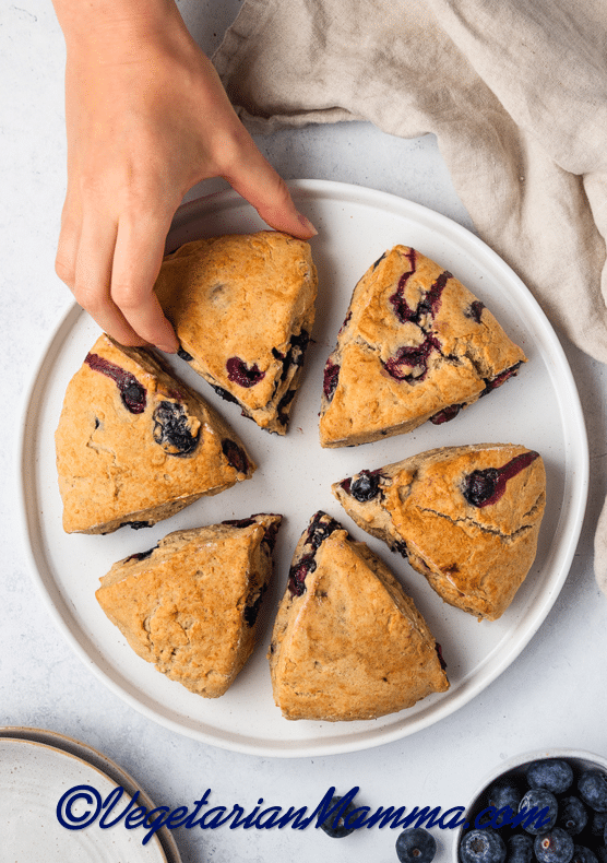 A round white plate of 6 scones on a table. A hand is picking one of them up.