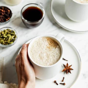 foamy creamy liquid in white cup on a white plate with a handing holding the cup from the left. On white plate is spices and a silver spoon. In the upper right hand corner is another white up filled with foam on a white plate.
