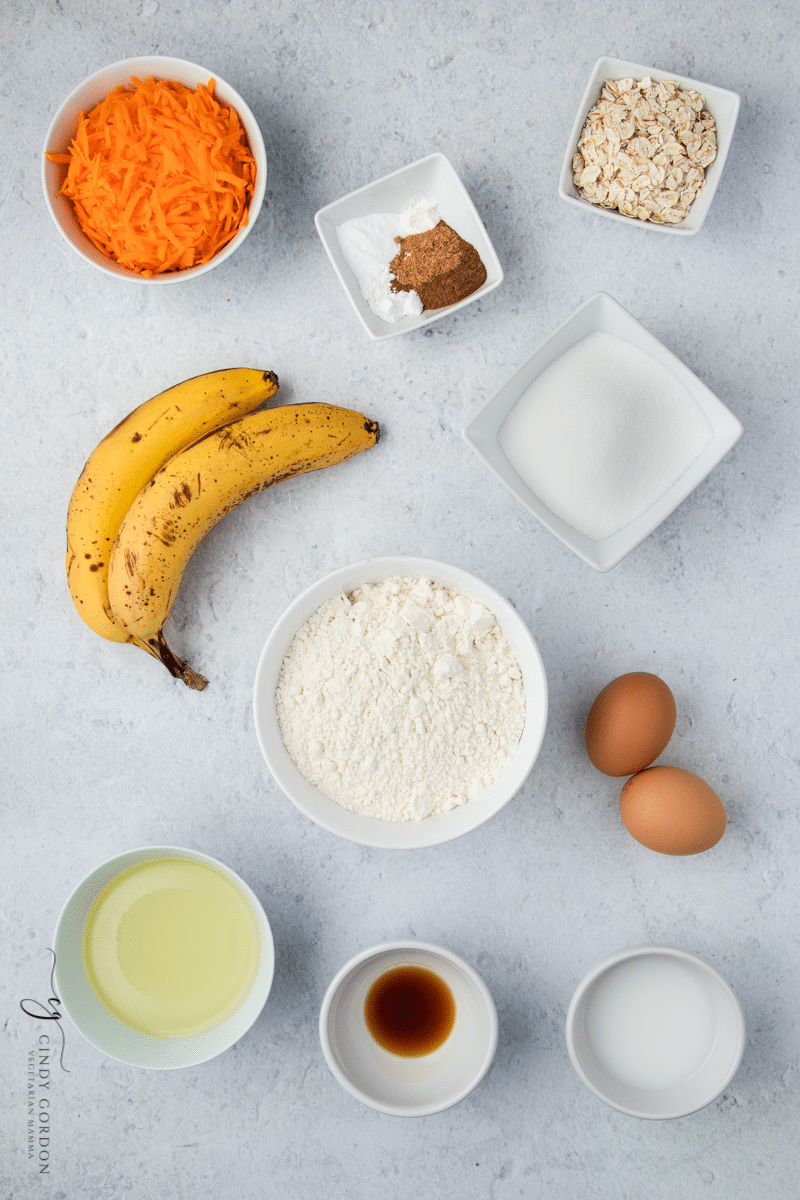 ingredients for banana carrot muffins, two ripe bananas, shredded carrot, two brown eggs and white bowls filled with liquids and powders