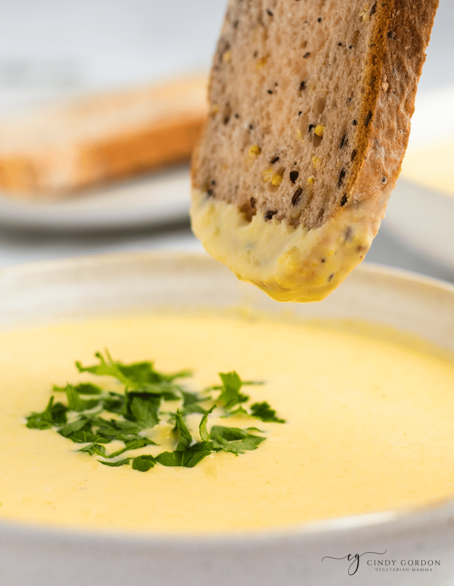 a bowl of corn potage with a slice of whole grain bread dipping into it.