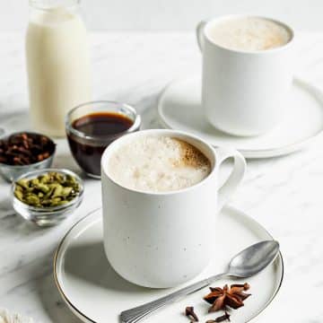 white foamy liquid in white mug on a white plate with a spoon and spices on top. In the back are clear small bowls of spices and maple syrup and a glass jar of milk.