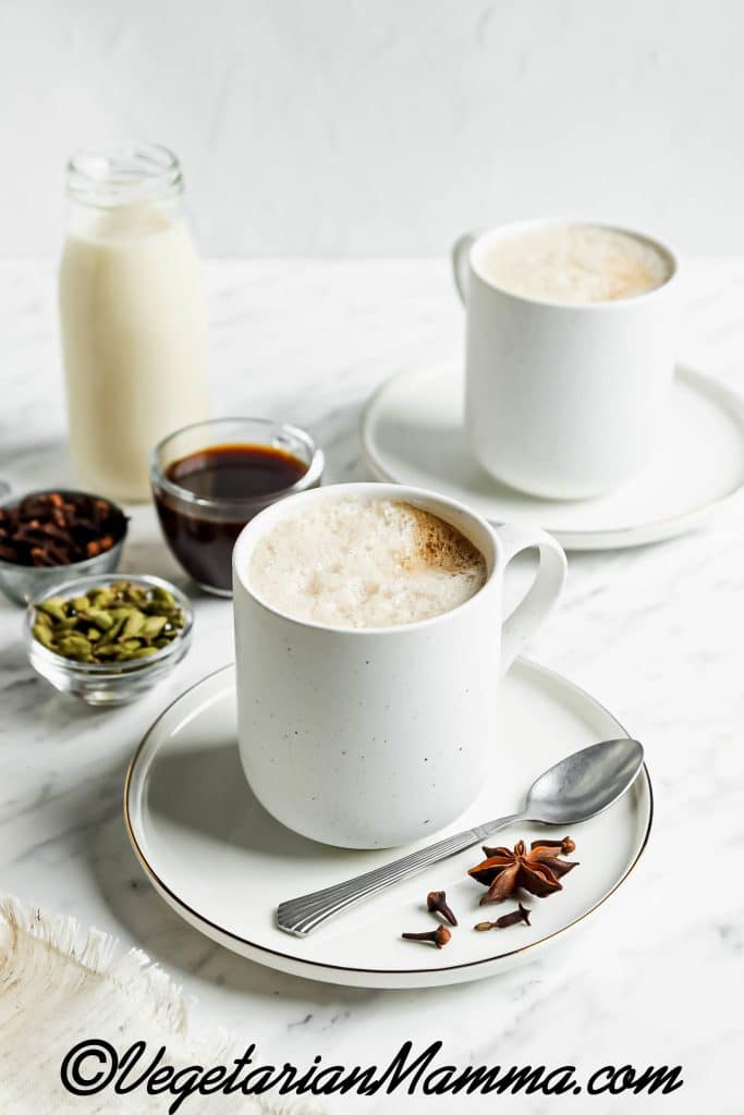 white foamy liquid in white mug on a white plate with a spoon and spices on top. In the back are clear small bowls of spices and maple syrup and a glass jar of milk.