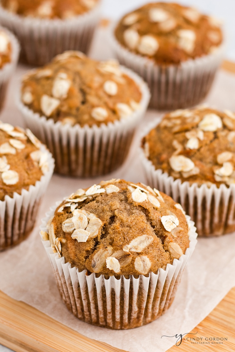 Up close angle of banana carrot muffins brown Muffins with oats on top in white cupcake liners sitting on white parchment paper