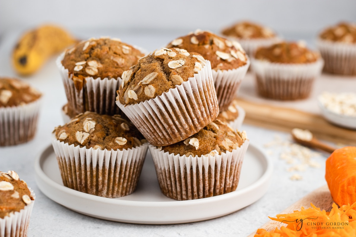 brown muffins in white liners on a white plate, stacked 2. high. Carrots on bottom right corner. On top of white background