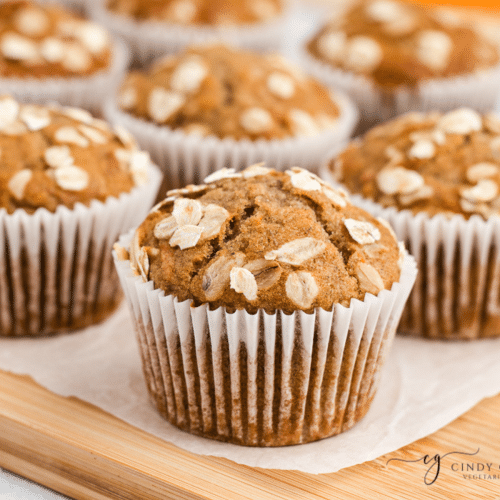 Up close angle of banana carrot muffins brown Muffins with oats on top in white cupcake liners sitting on white parchment paper