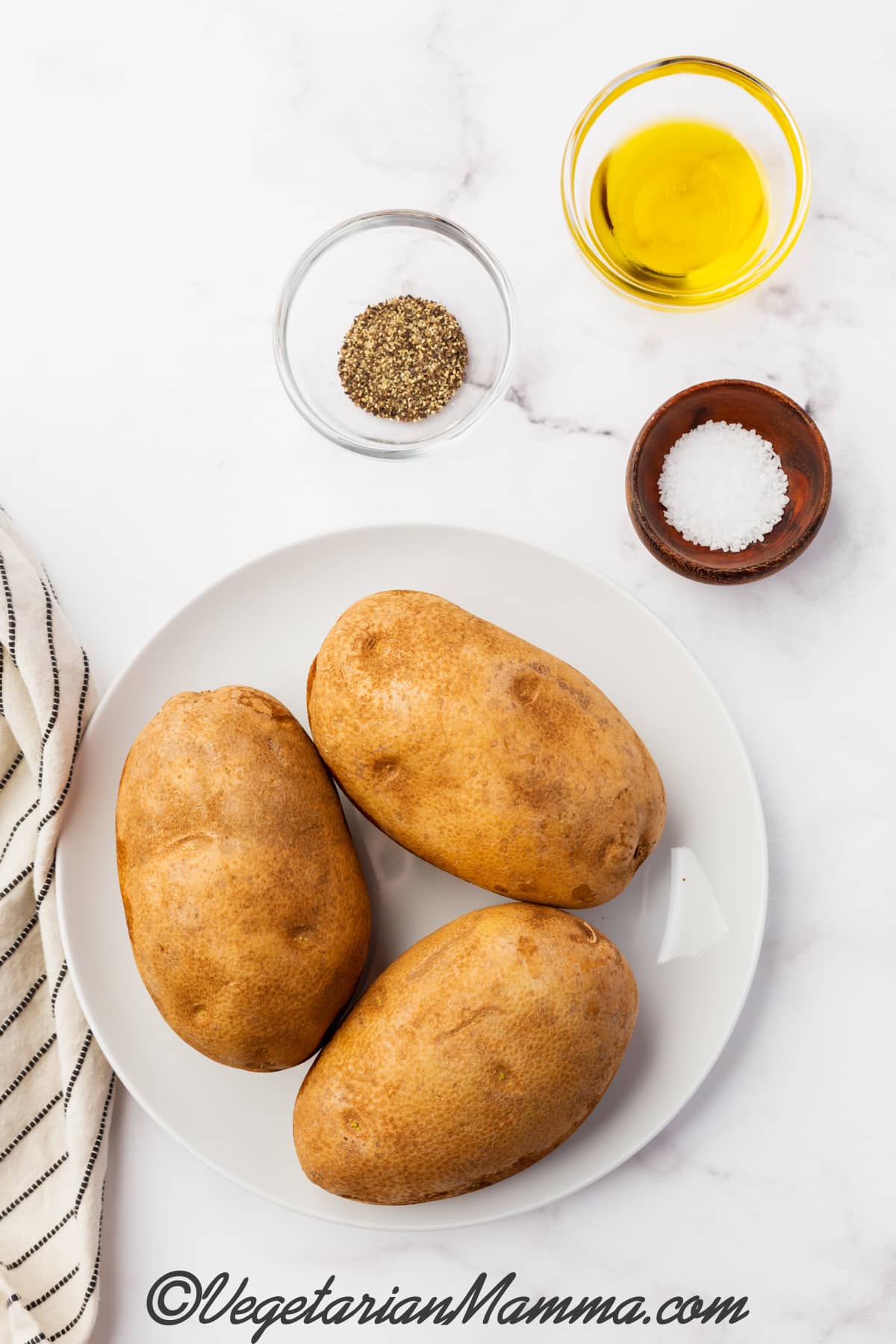 three uncooked brown potatoes in white plate, salt in a bowl, seasoning in a bowl and oil in a bowl