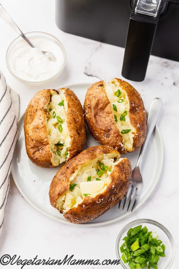three air fryer baked potatoes with butter and chives with a bowl of sour cream.