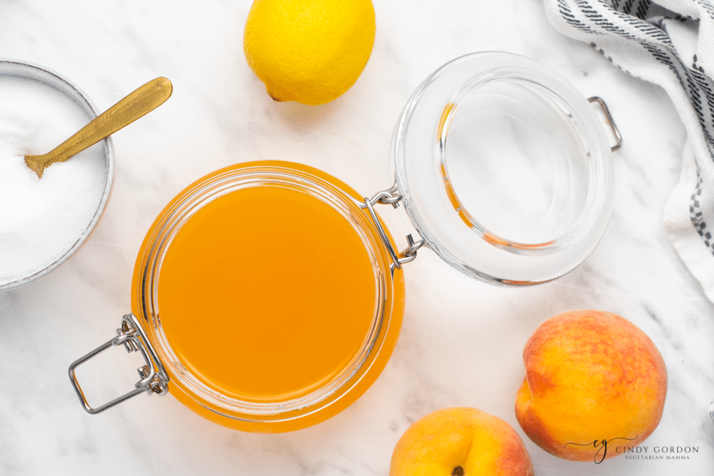 view from above of a glass bail jar filled with peach simple syrup with the lid open. There are two peaches, a lemon, and a bowl of sugar nearby