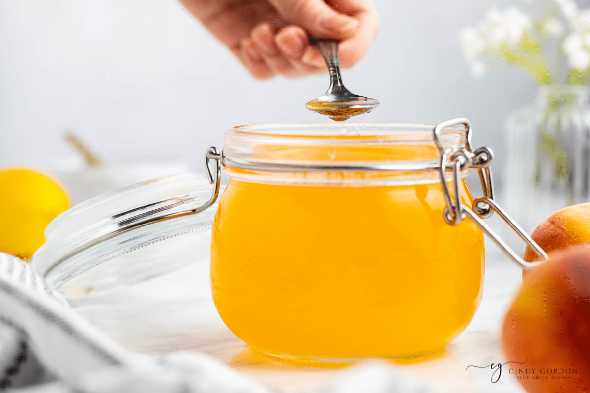 a jar of peach simple syrup. a hand is holding a spoon above it, scooping some out of the jar. 