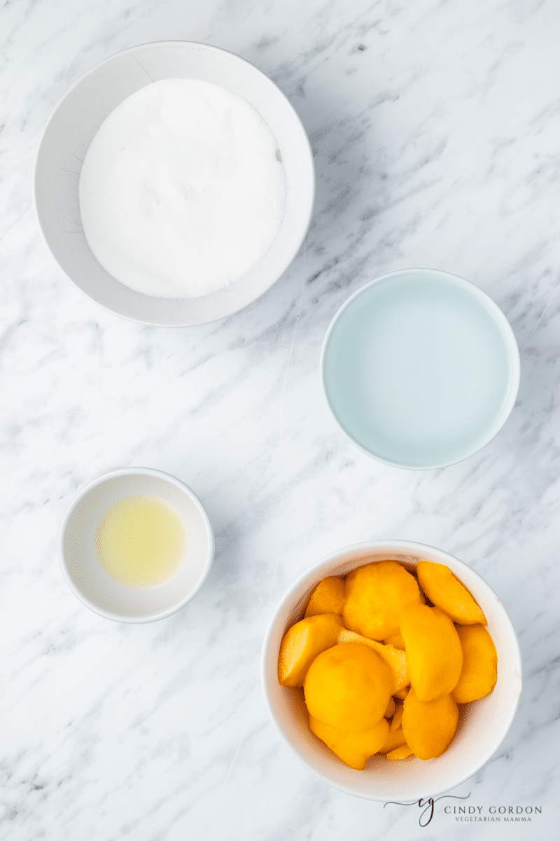 viewed from above, a bowl of peeled and sliced peaches, a bowl of sugar, a small bowl of lemon juice, and a small bowl of water on a marble counter. The ingredients for peach simple syrup