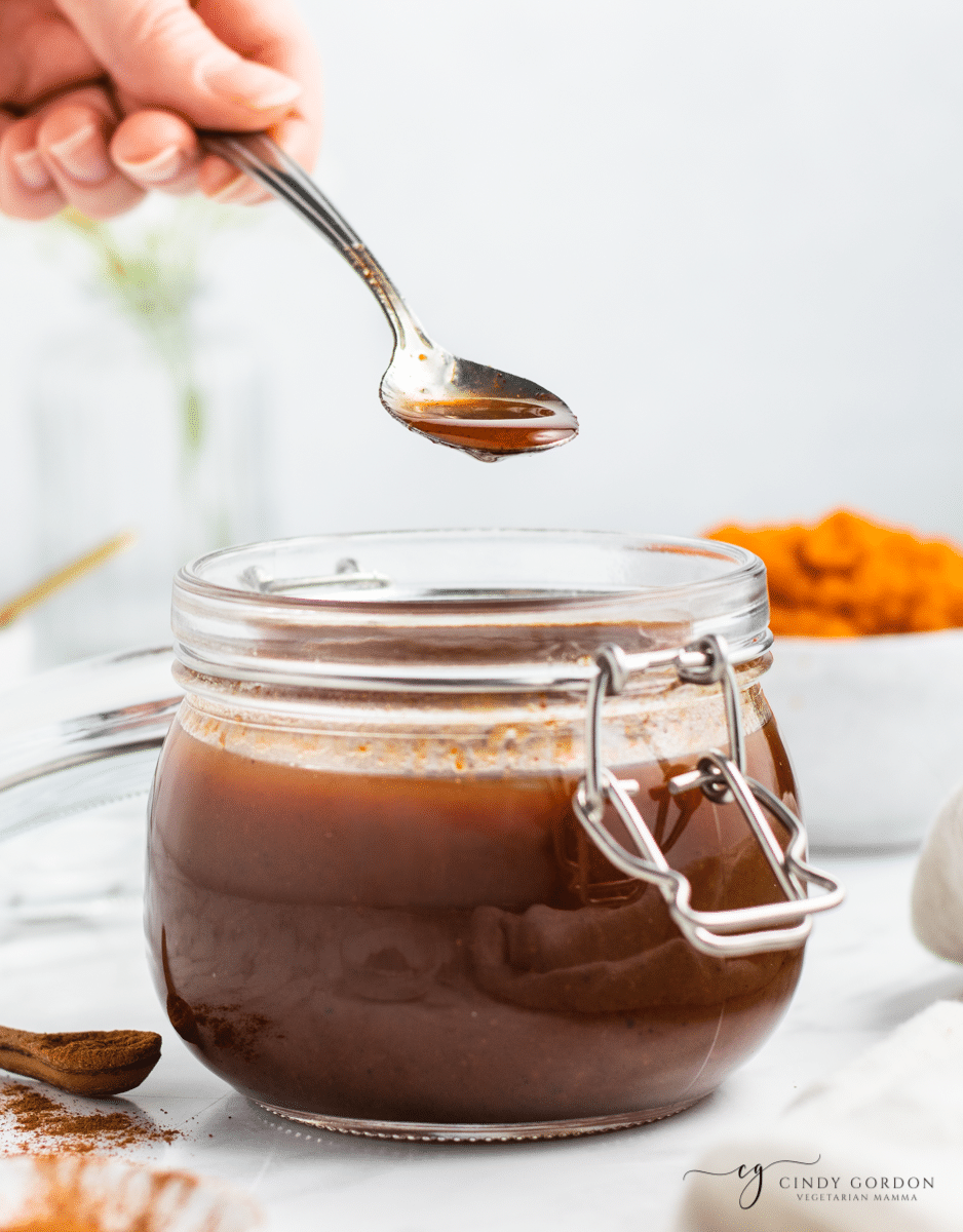 vertical photo of white backdrop. Brown liquid in glass jar with hand holding a spoon. Dripping from the spoon is sugar free pumpkin spice syrup