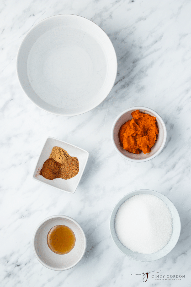 on a white background and white bowls, you will see the ingredients for sugar free pumpkin spice syrup. In the bowl are: spices, sugar substitute, pumpkin puree and a brown liquid