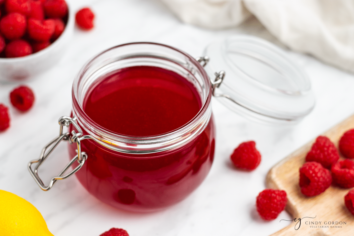 a jar of raspberry simple syrup on a counter, surrounded by fresh raspberries