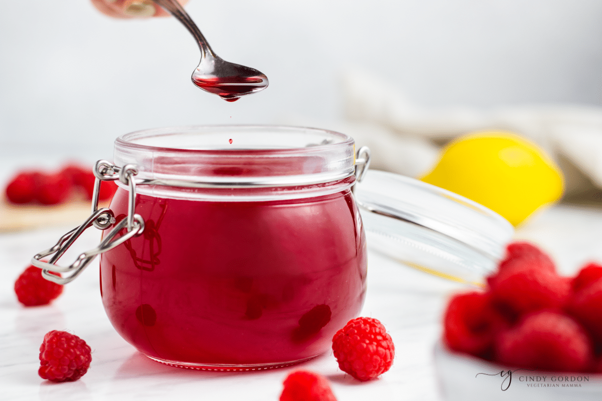 a rounded jar filled with raspberry syrup. A teaspoon is lifting some syrup out. 