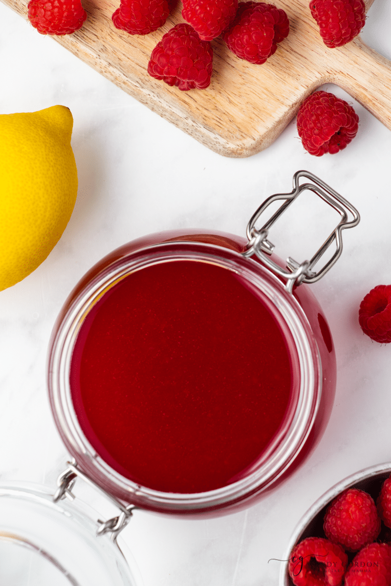 an open bail jar of raspberry simple syrup, viewed from above next to a cutting board of raspberries and a lemon