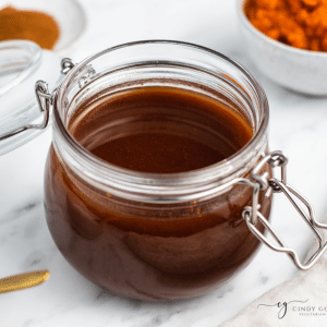 vertical photo, brown liquid in glass jar with latch lid off in center of photo. Wooden spoon with orange spice spilled at the bottom of the photo. White bowl with orange substance on top left of photo.