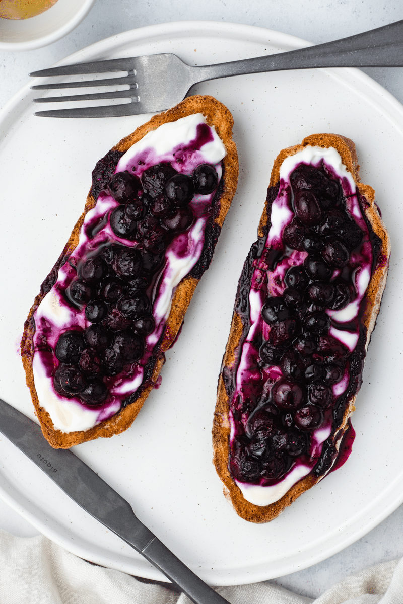Blueberry toast - brown bread slices, with white yogurt on top, then purple blueberry spread on top and honey drizzled on top. On a white plate, on a white background with a silver muted fork