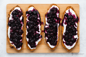 Blueberry toast - brown bread slices, with white yogurt on top, then purple blueberry spread on top and honey drizzled on top. On a white plate, on a wooden cutting board
