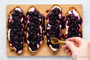 Blueberry toast - brown bread slices, with white yogurt on top, then purple blueberry spread on top and honey drizzled on top. On a white plate, on a wooden cutting board - hand using a gold spoon putting honey on bread slices