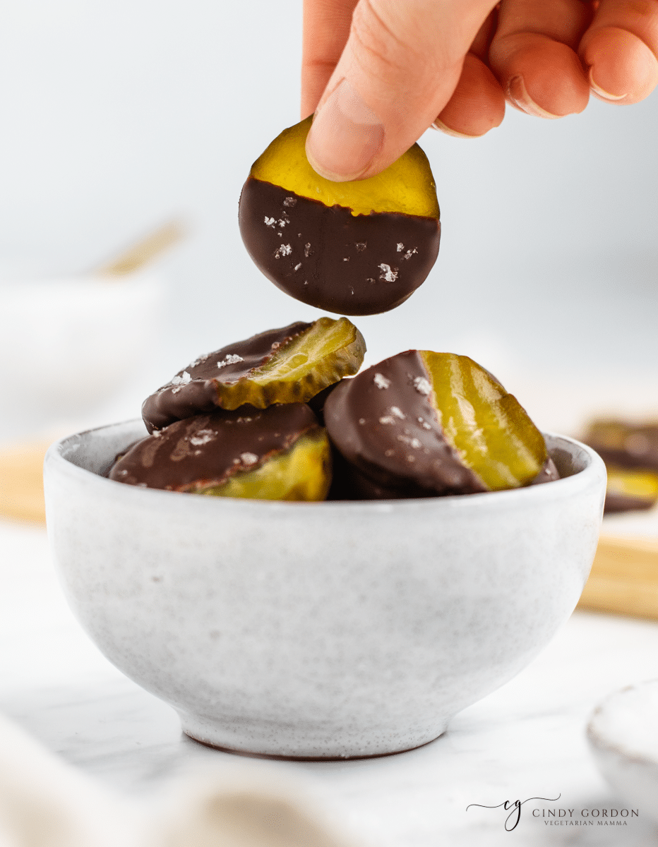 green pickle circles with half pickle showing, other half dipped into dark brown chocolate with white sea salt flakes ina white bowl with a hand picking up one pickle circle