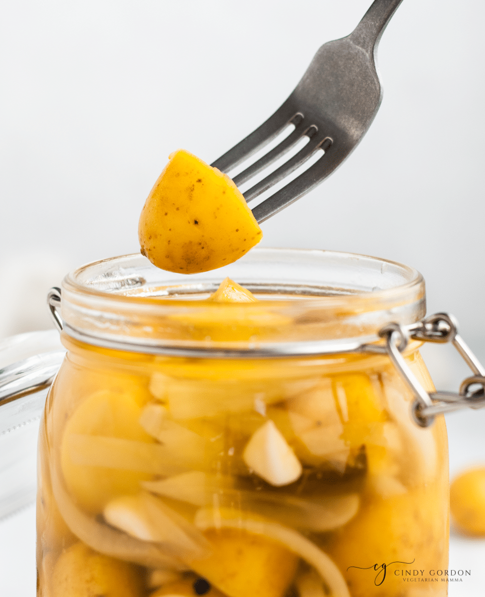 vertical imae of clear glass jar with yellow baby round potatoes cut in half with thin white onion slices in milkly liquid. a fork above the open jar with one potato on it