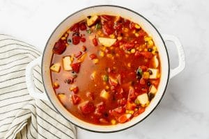 red liquid in white dutch oven with corn and diced tomatoes visable