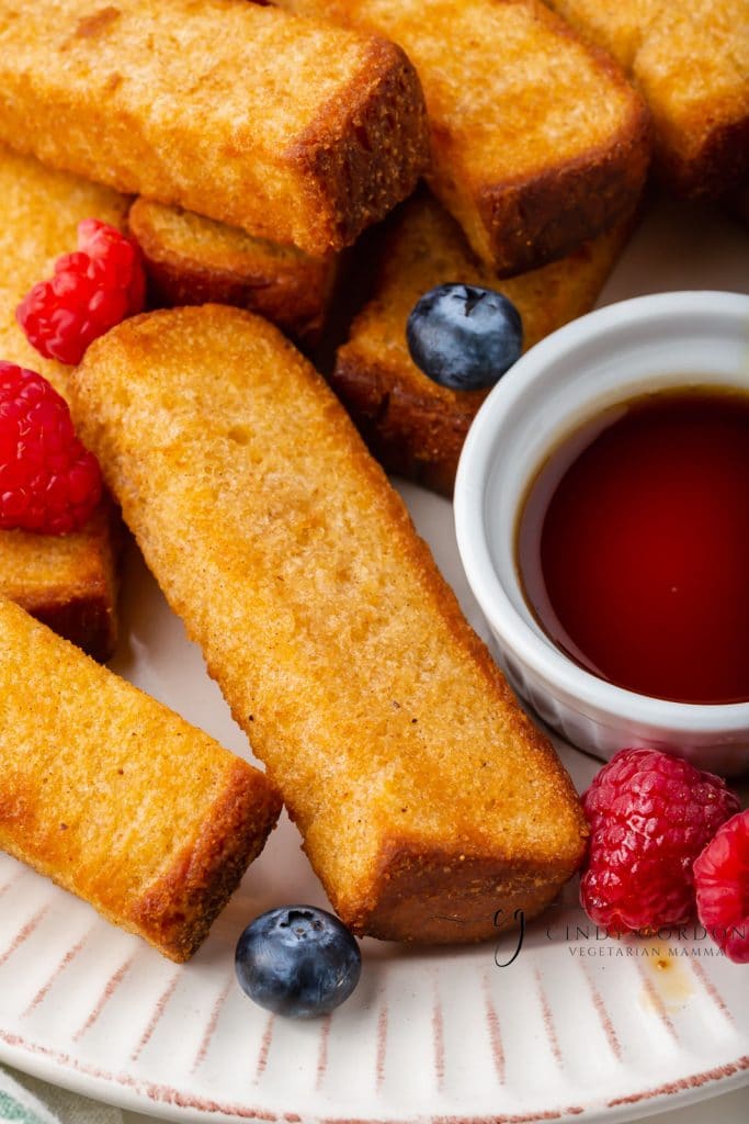 golden brown air fryer french toast sticks on a white plate with blueberries and raspberries sprinkled on top and a white little cup of brown liquid