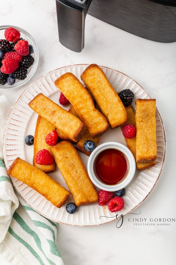 golden brown air fryer french toast sticks on a white plate with blueberries and raspberries sprinkled on top and a white little cup of brown liquid