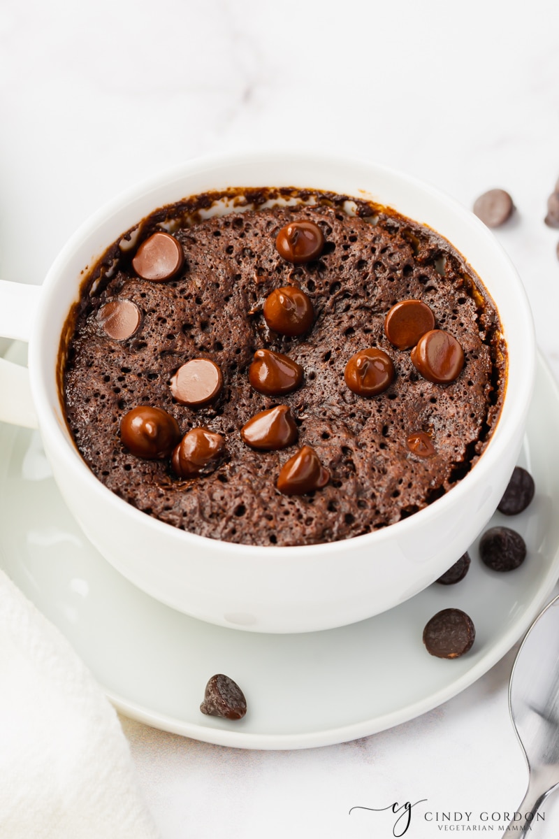 Pictured is a vegan mug brownie, in a white large coffee cup is a fluffy brown cooked batter with shiny chocolate chip morsels on top. Cup is sitting on a white plate and some chocolate chips have been spilled onto the plate