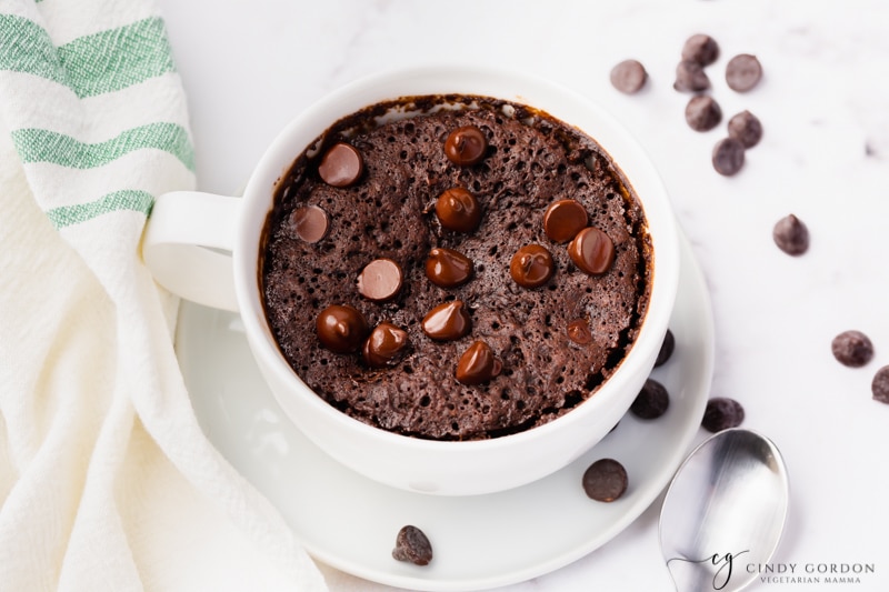 Pictured is a vegan mug brownie, in a white large coffee cup is a fluffy brown cooked batter with shiny chocolate chip morsels on top. Cup is sitting on a white plate and some chocolate chips have been spilled onto the plate