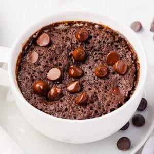 Pictured is a vegan mug brownie, in a white large coffee cup is a fluffy brown cooked batter with shiny chocolate chip morsels on top. Cup is sitting on a white plate and some chocolate chips have been spilled onto the plate