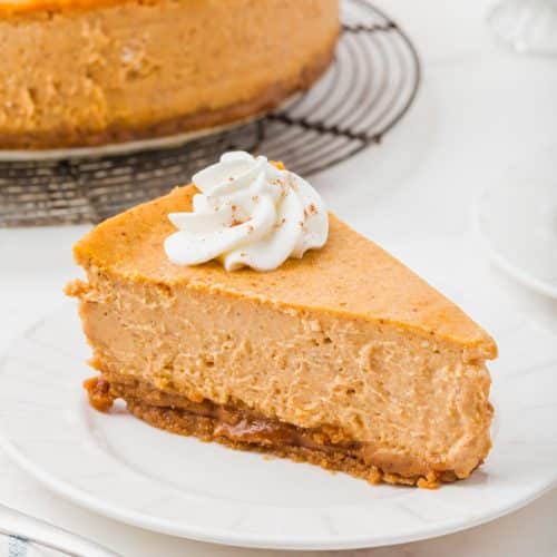 A slice of pumpkin cheesecake on a white plate with a fork at the ready. The rest of the cake is in the background, waiting to be devoured.