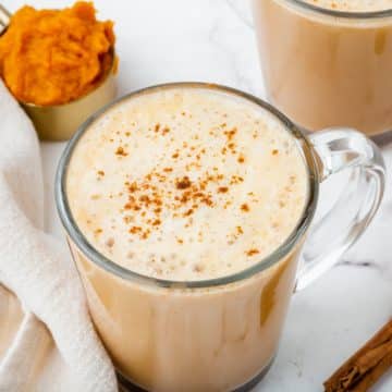 A frothy pumpkin spiced latte in a clear coffee mug surrounded by cinnamon and pumpkin puree