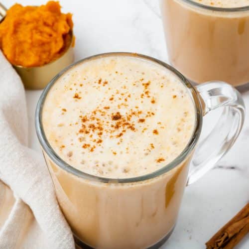 A frothy pumpkin spiced latte in a clear coffee mug surrounded by cinnamon and pumpkin puree