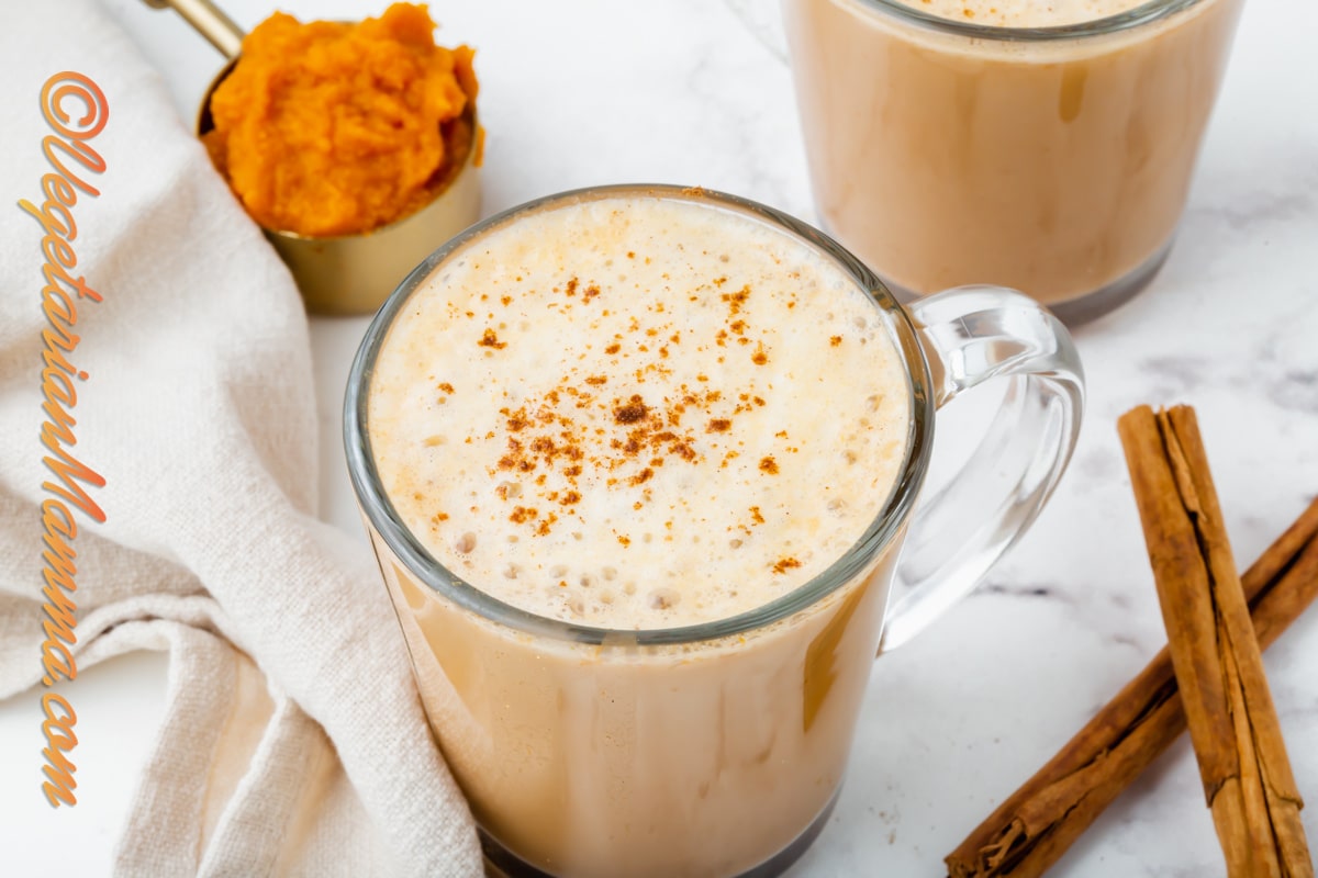 A frothy pumpkin spiced latte in a clear coffee mug surrounded by cinnamon and pumpkin puree