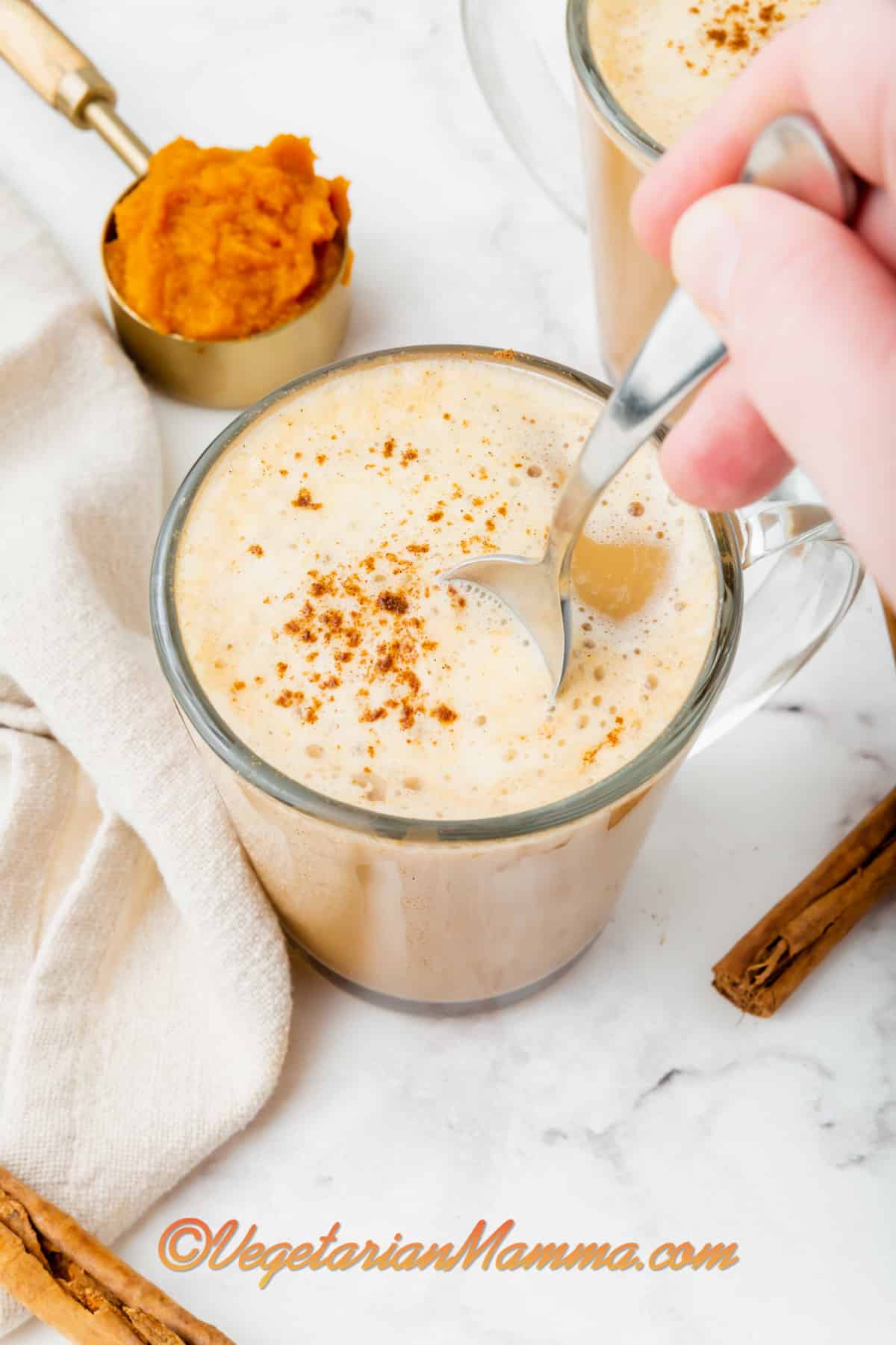 Someone stirring a pumpkin spice latte with a silver spoon in a clear mug