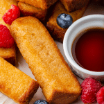 golden brown air fryer french toast sticks on a white plate with blueberries and raspberries sprinkled on top and a white little cup of brown liquid