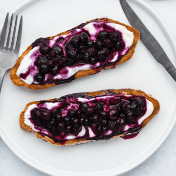 Blueberry toast - brown bread slices, with white yogurt on top, then purple blueberry spread on top and honey drizzled on top. On a white plate, on a white background with a silver muted fork and knife