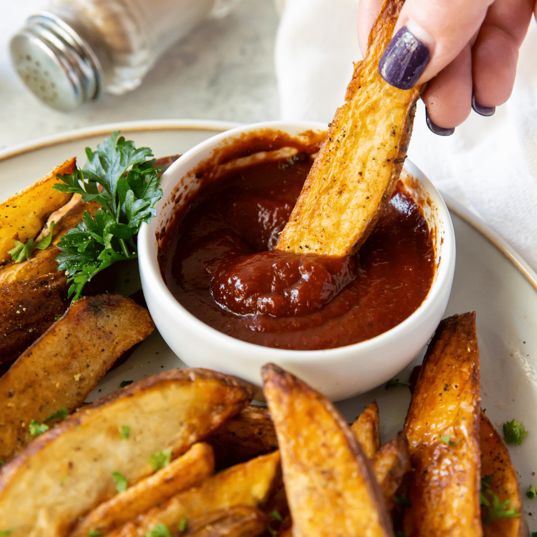 finger with purple nail dipping a single air fryer potato wedge into ketchup on top of a plate full of cooked and seasoned air fryer potato wedges